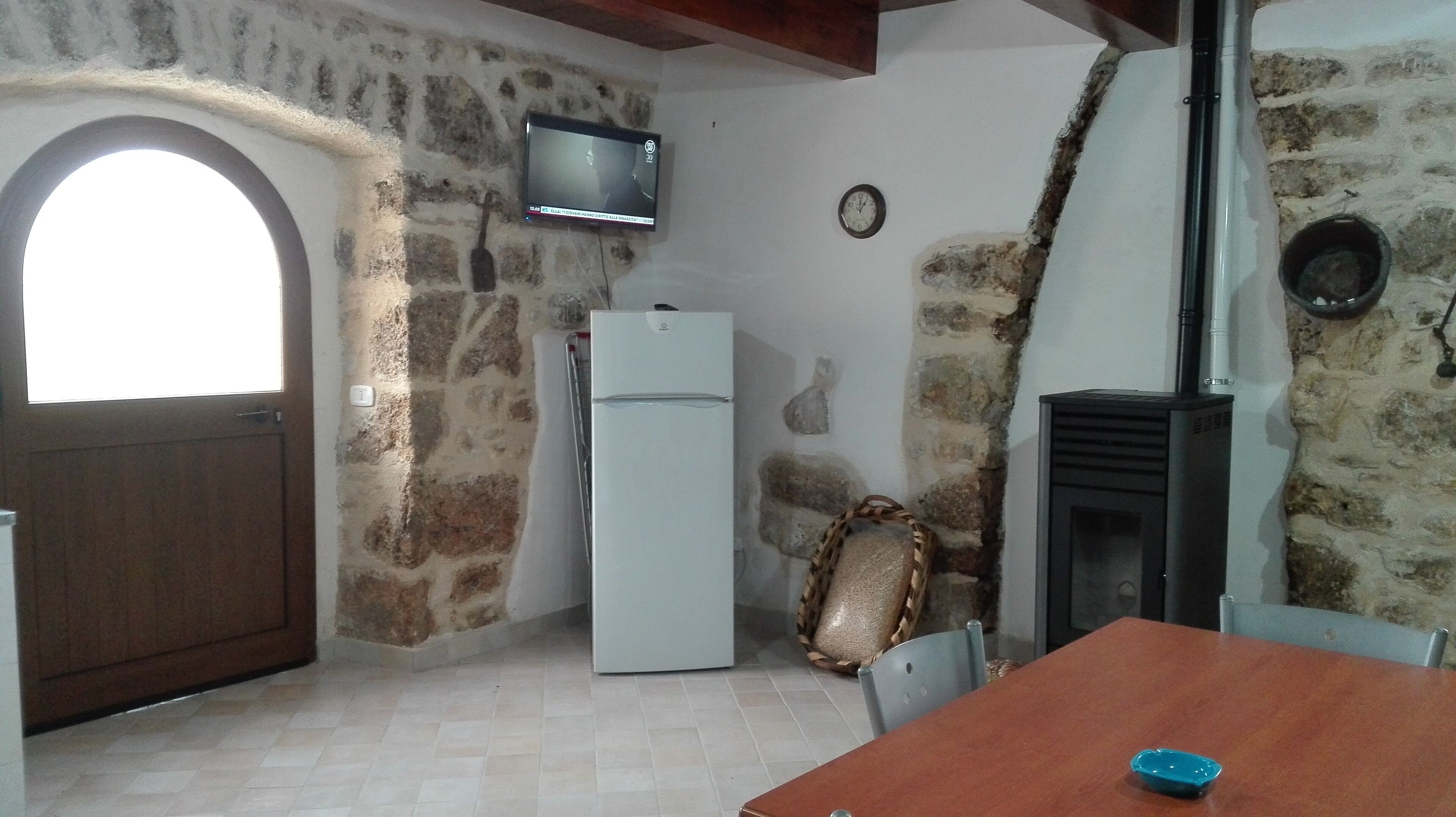 Una habitación rústica con pared de piedra, puerta de madera, refrigerador blanco, televisor montado en la pared, un reloj y chimenea.