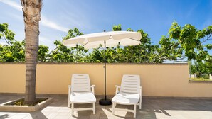 Two white lounge chairs under a large white umbrella, positioned on a tiled patio with a palm tree and a beige wall in the background. - Newly built villa: Car garage, 3 bedrooms, 6p let, large kitchen, gardens (Tuglie)