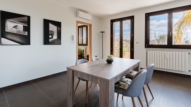 A modern dining area with a wooden table and chairs, framed artwork on the walls, and large windows with a view of the outdoors. - Newly built villa: Car garage, 3 bedrooms, 6p let, large kitchen, gardens (Tuglie)