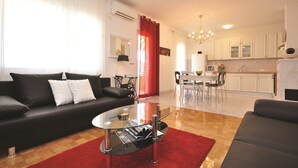 Una sala de estar moderna con un sofá negro, una mesa de centro de vidrio y una alfombra roja. Se observa una zona de cocina con armarios blancos y una mesa de comedor con sillas.