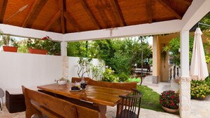 Un porche cubierto con una mesa y bancos de madera, rodeado de suelo de piedra y plantas en macetas.