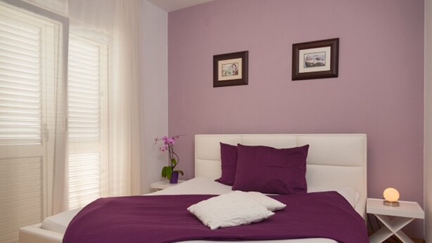 Un dormitorio con una cama blanca, ropa de cama morada, una mesita de noche con una lámpara y dos cuadros en la pared.