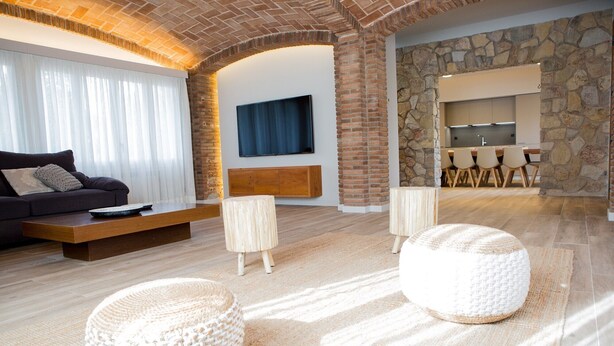 A modern living room with a sofa, coffee table, and two poufs. A stone wall and a kitchen area are visible in the background.