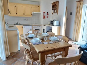 A compact kitchen with modern appliances, a dining area with a wooden table set for four, and a view of the outdoors through a window. - No 3 Ballyconneely Cottage, Aillebrack (Ballyconneely)
