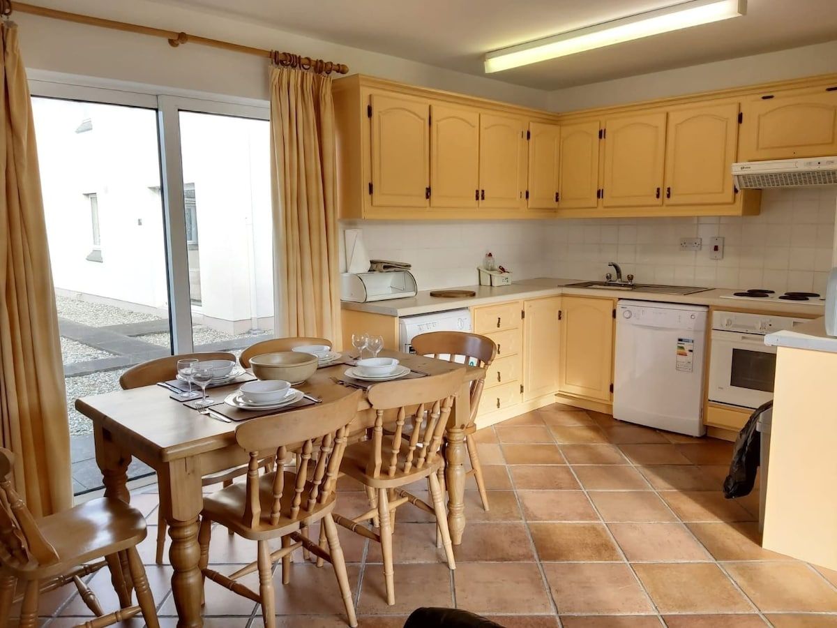 A kitchen with wooden cabinets, a dining table with chairs, a window with curtains, and a tiled floor.