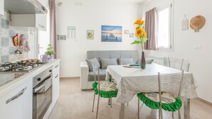 A compact kitchen with a gas stove, oven, and white cabinets. A grey sofa and a dining table with chairs are in the dining area. - Salento Rosa dei Venti - Scirocco (Surbo)