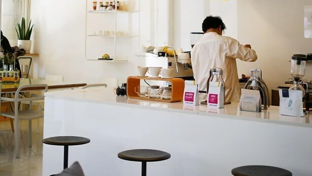 A modern kitchen with a bar area, featuring a person preparing drinks, a coffee machine, and a shelf with kitchenware. - Concept Yard Chiangrai - Adults Only - Hostel (Chiang Rai)