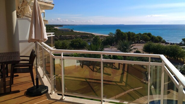 A balcony with a clear view of the sea and beach, a glass railing, and a table with chairs.