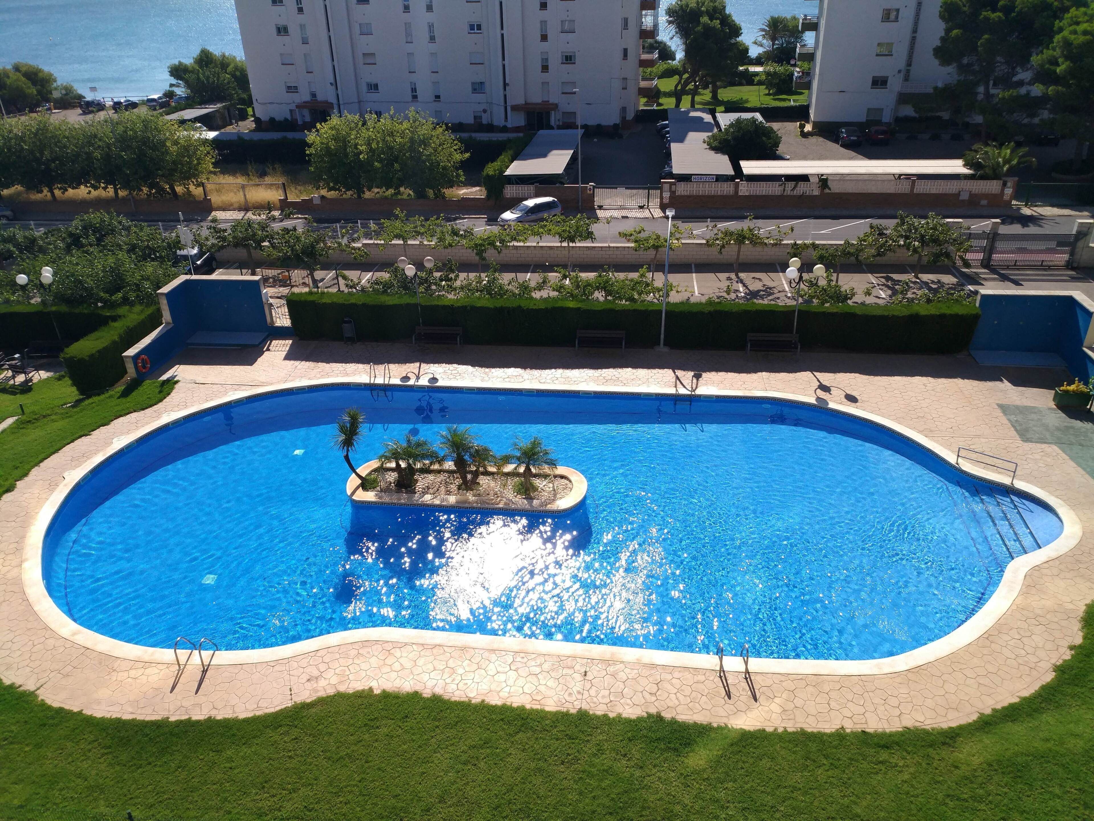 Ein Schwimmbecken mit einer kleinen Insel, umgeben von einer gepflasterten Terrasse und Rasen.