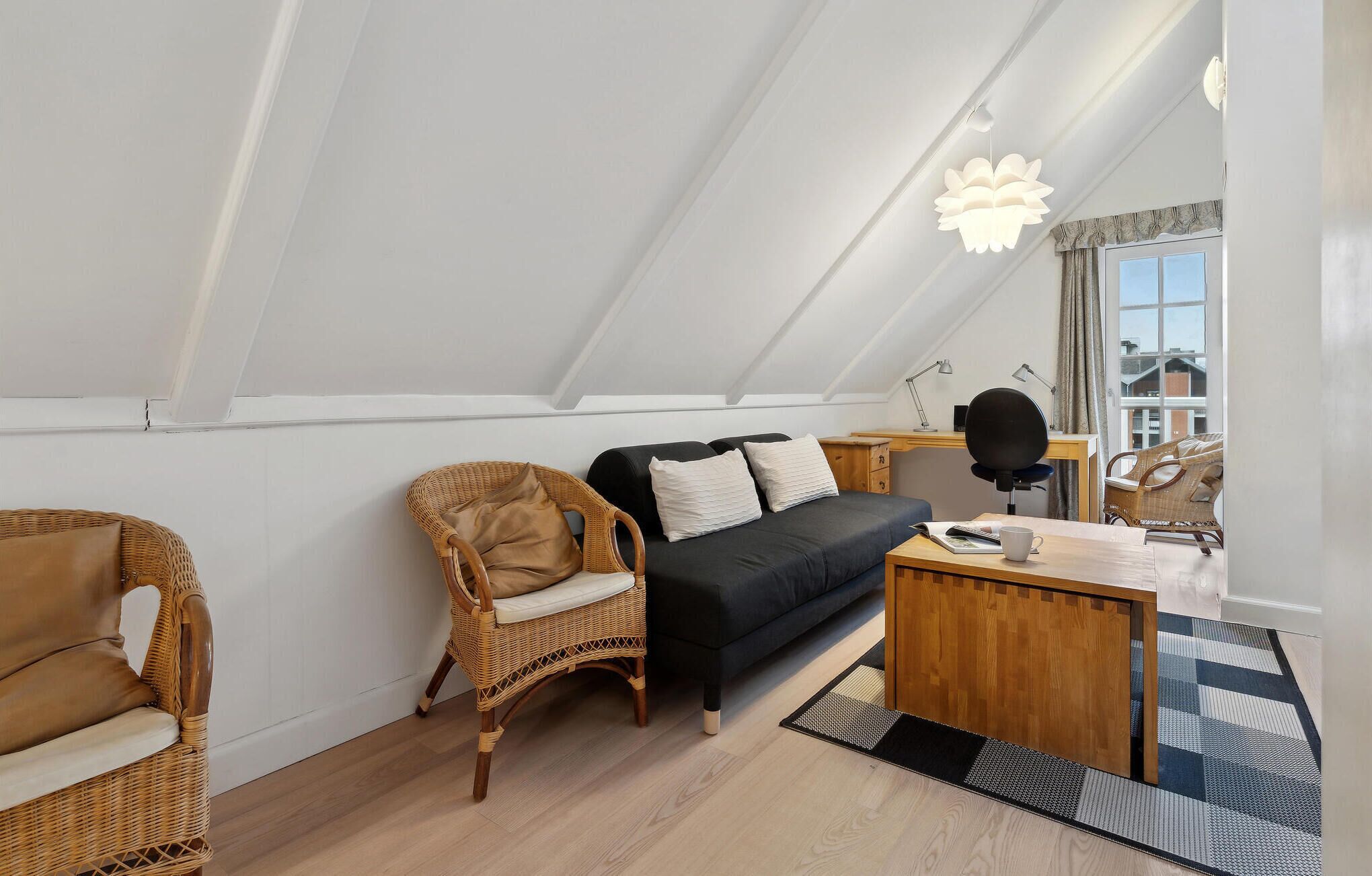 Un acogedor dormitorio tipo loft con un sofá negro, sillas de mimbre, un escritorio de madera y un ventanal con vistas a la ciudad.