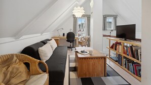 A cozy loft-style living space with a black sofa, a wooden coffee table, a wicker chair, a bookshelf, and a TV.