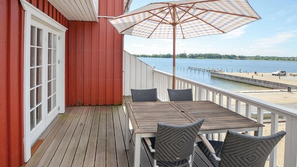 A wooden deck with a table and chairs, a checkered umbrella, and a view of a lake and dock.