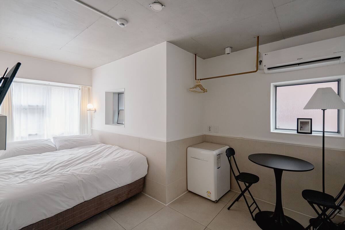 A hotel room with a bed, a small table with chairs, a compact refrigerator, a wall-mounted air conditioner, and a window with a lamp.