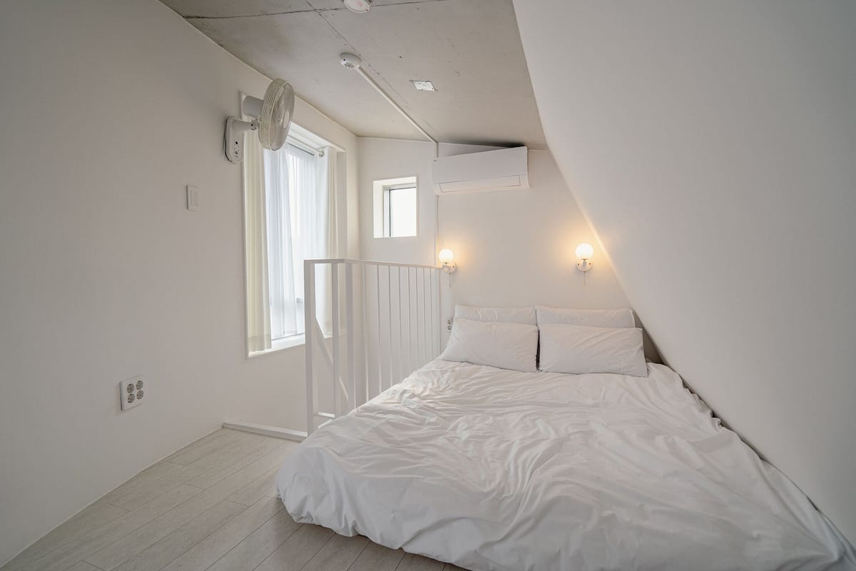 A bedroom with a sloped ceiling, a bed with white bedding, a window with curtains, and wall-mounted lighting.