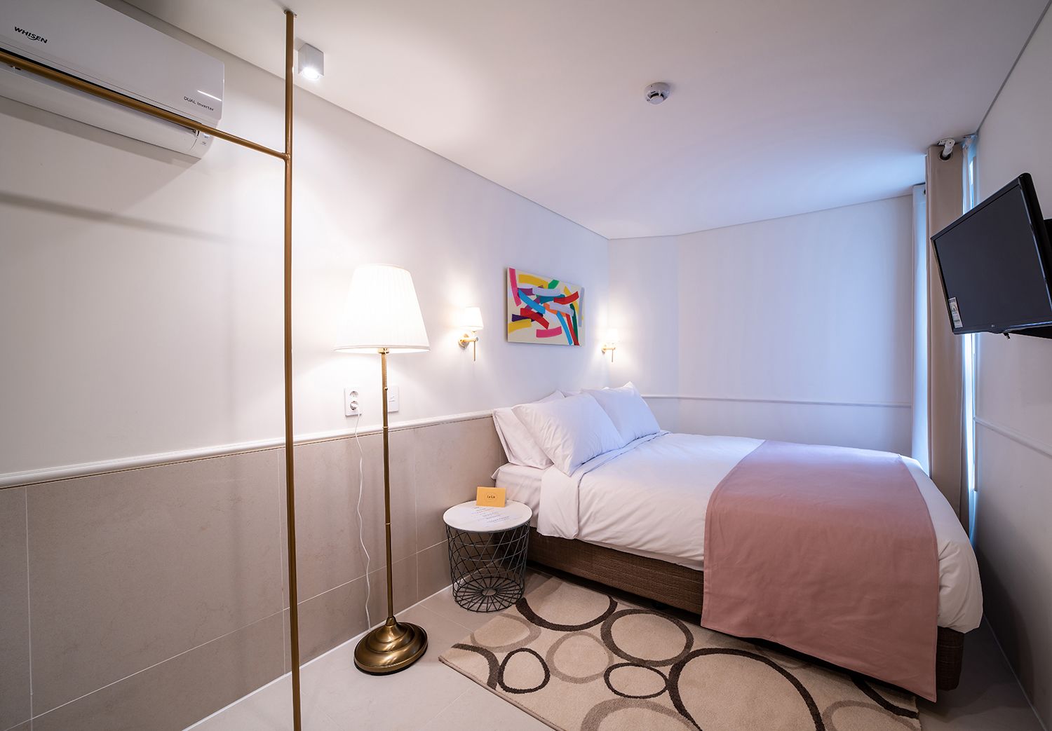 A modern hotel room with a bed, a bedside table with a lamp, a small round table with a card, a colorful abstract painting on the wall, and a mounted TV.