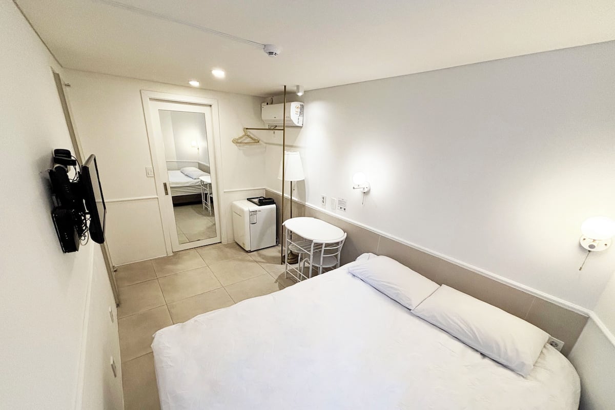 A hotel room with a bed, a small table, a chair, a mirror, a wardrobe, and a wall-mounted heater.