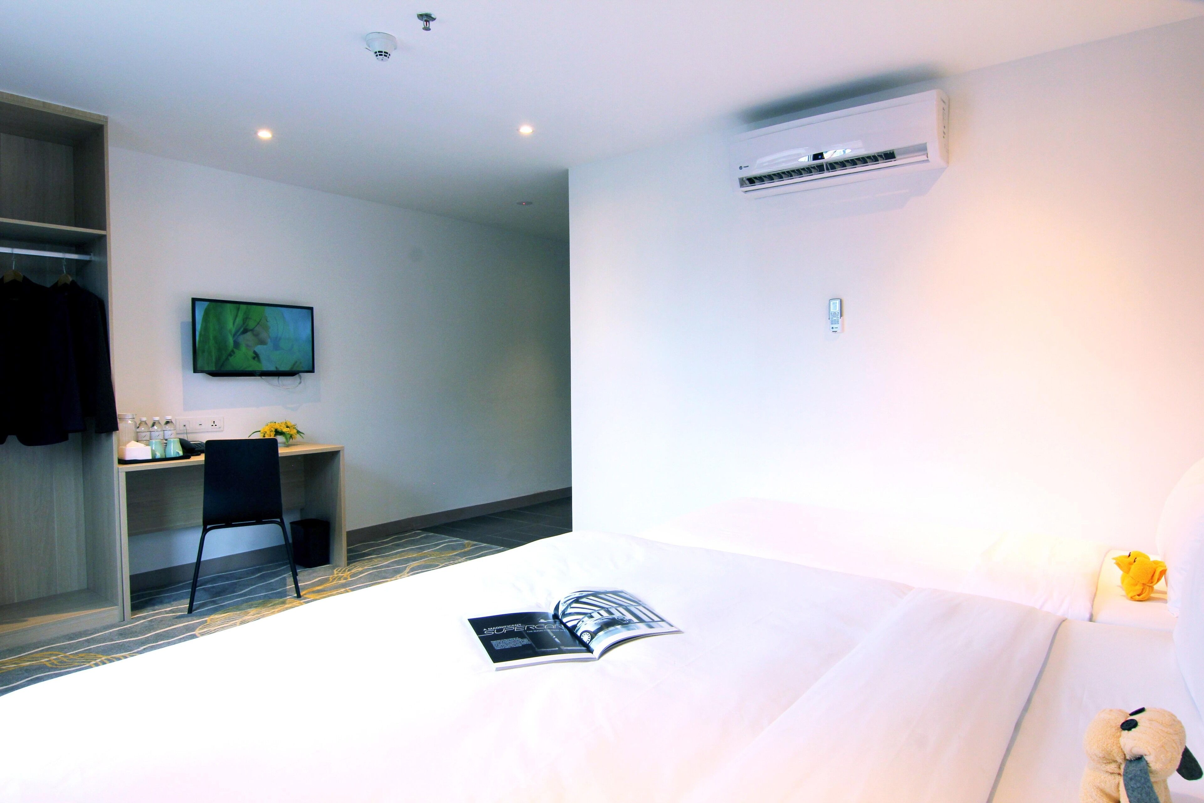 A hotel room with a bed, a desk with a chair, a television on the wall, and an air conditioning unit.