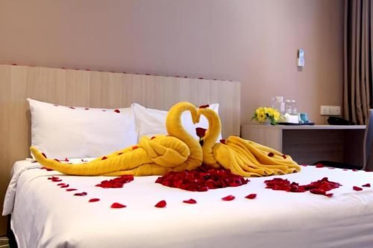 A hotel room with a bed adorned with a heart-shaped towel arrangement and scattered rose petals.
