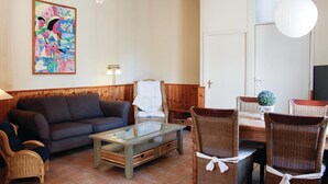 A living room with a sofa, a glass-top coffee table, wicker chairs, a dining table, and a colorful painting on the wall.