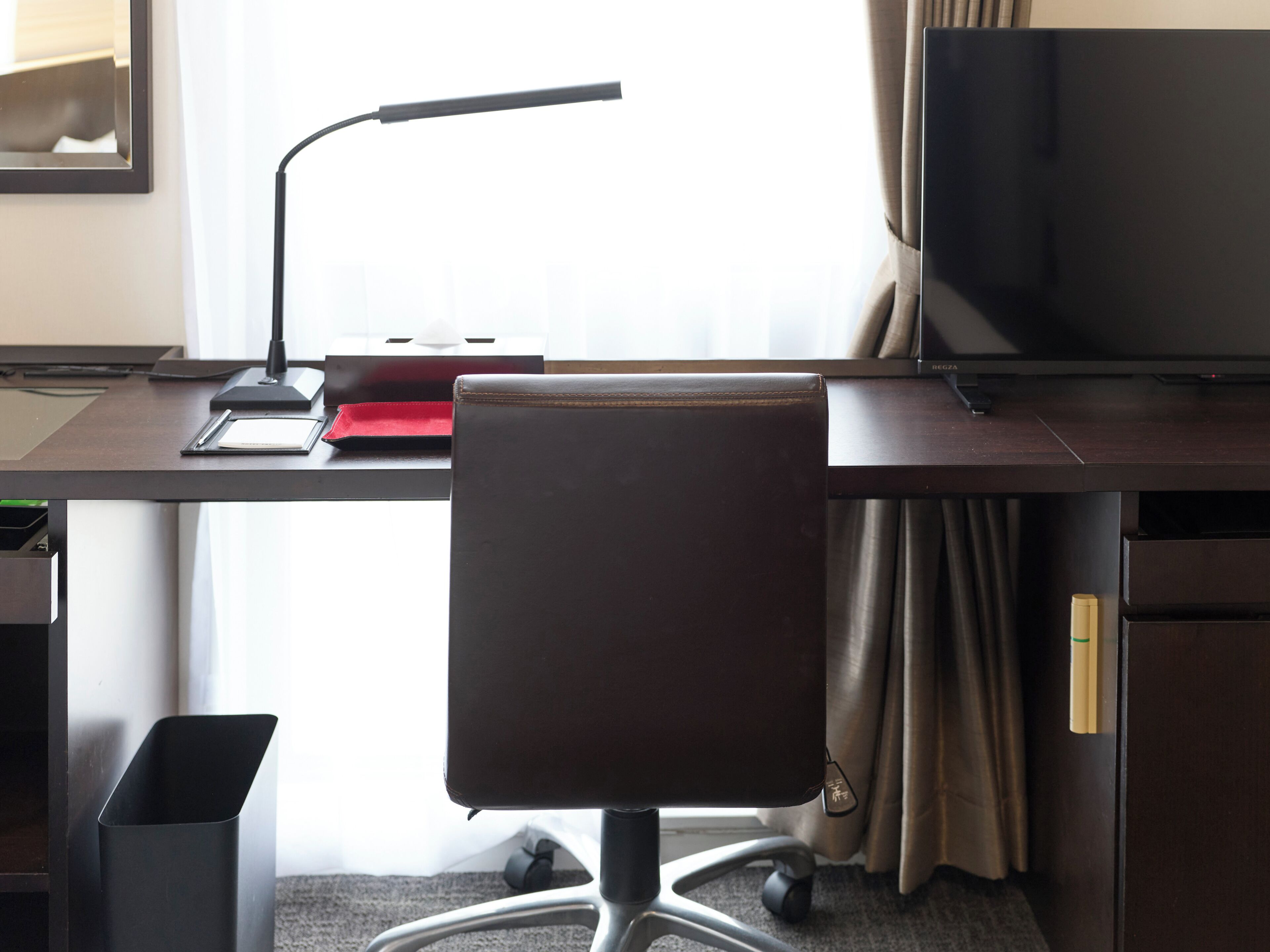 Een hotelkamer met een bureau, een stoel, een prullenbak, een lamp, een televisie en een raam met gordijnen.