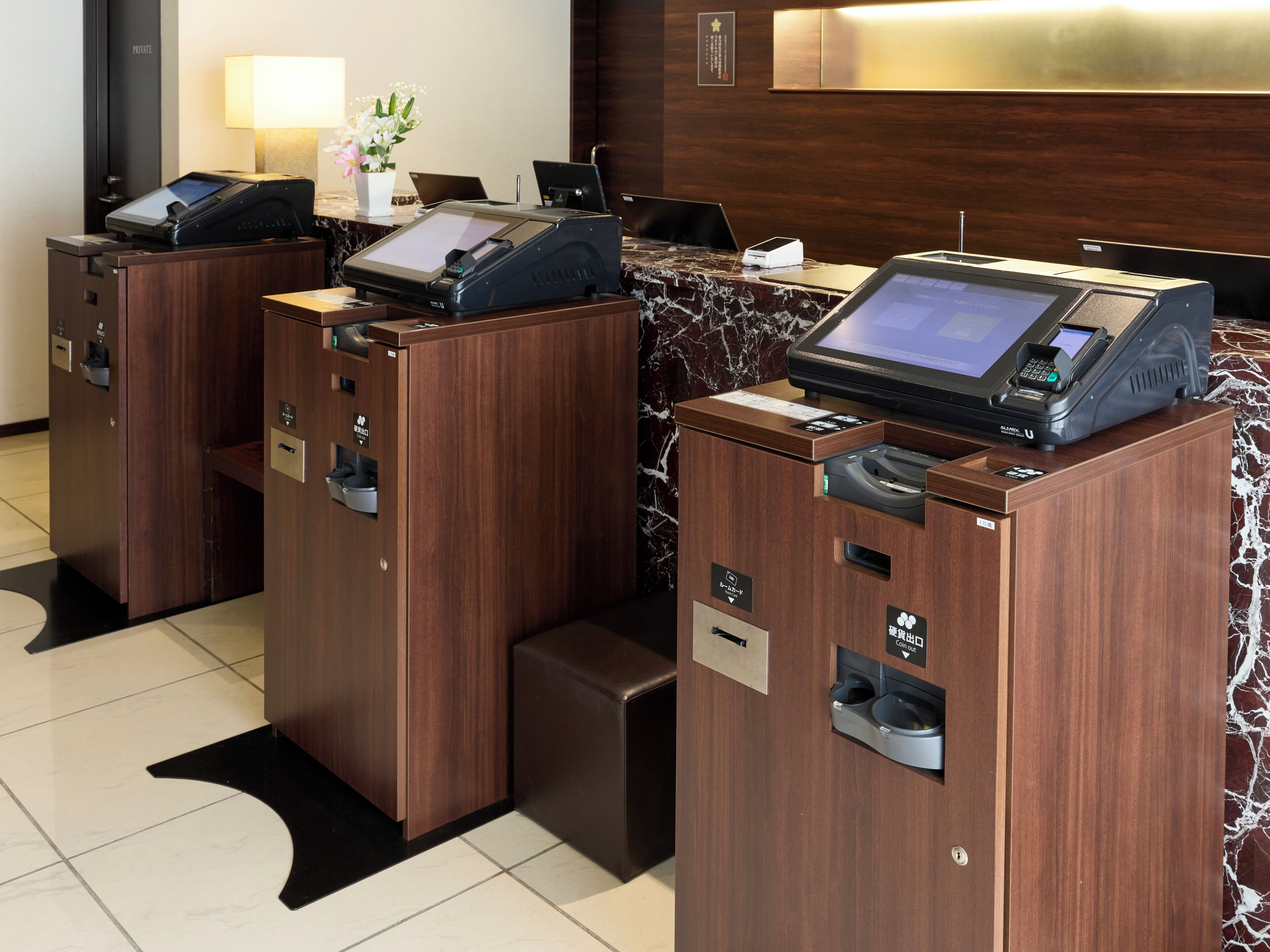 A hotel reception area with two self-service kiosks, a reception desk, and a wall-mounted sign.