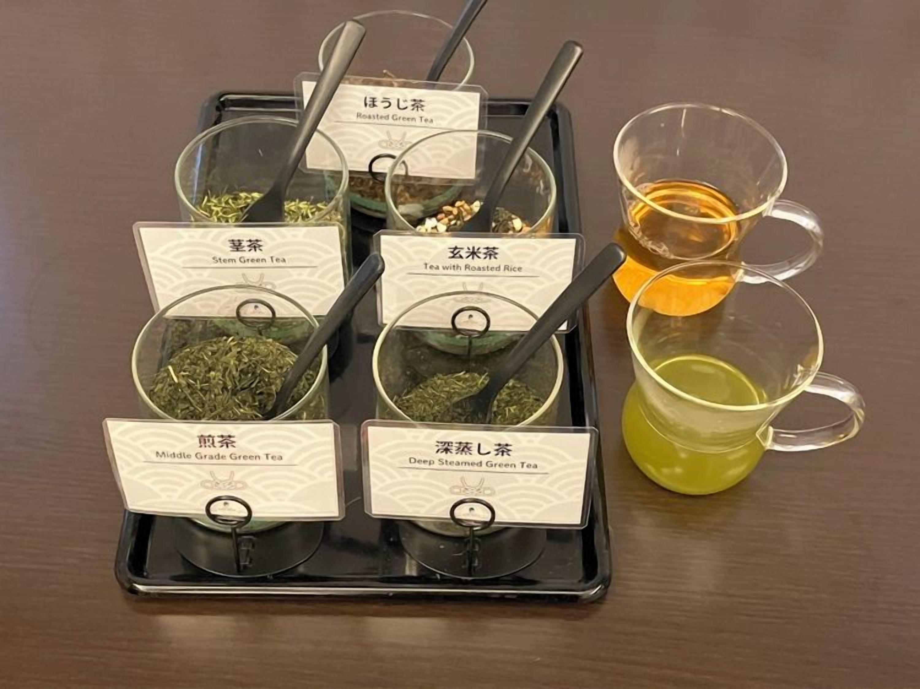 A tray with six labeled containers of tea, each with a spoon, and two cups of tea on the side.