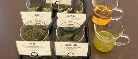 A tray with six labeled containers of tea, each with a spoon, and two cups of tea on the side.