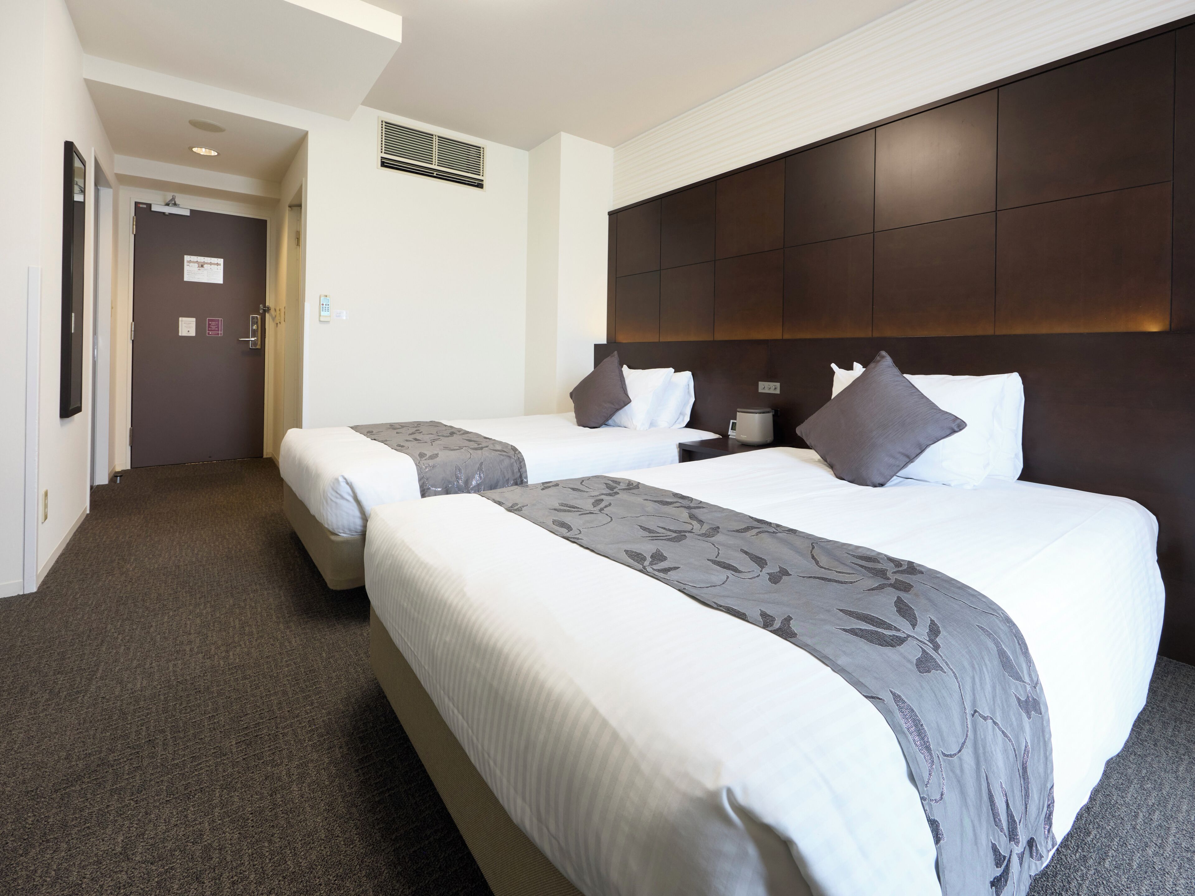 A hotel room with two beds, a dark headboard, a small bedside table, and a carpeted floor.