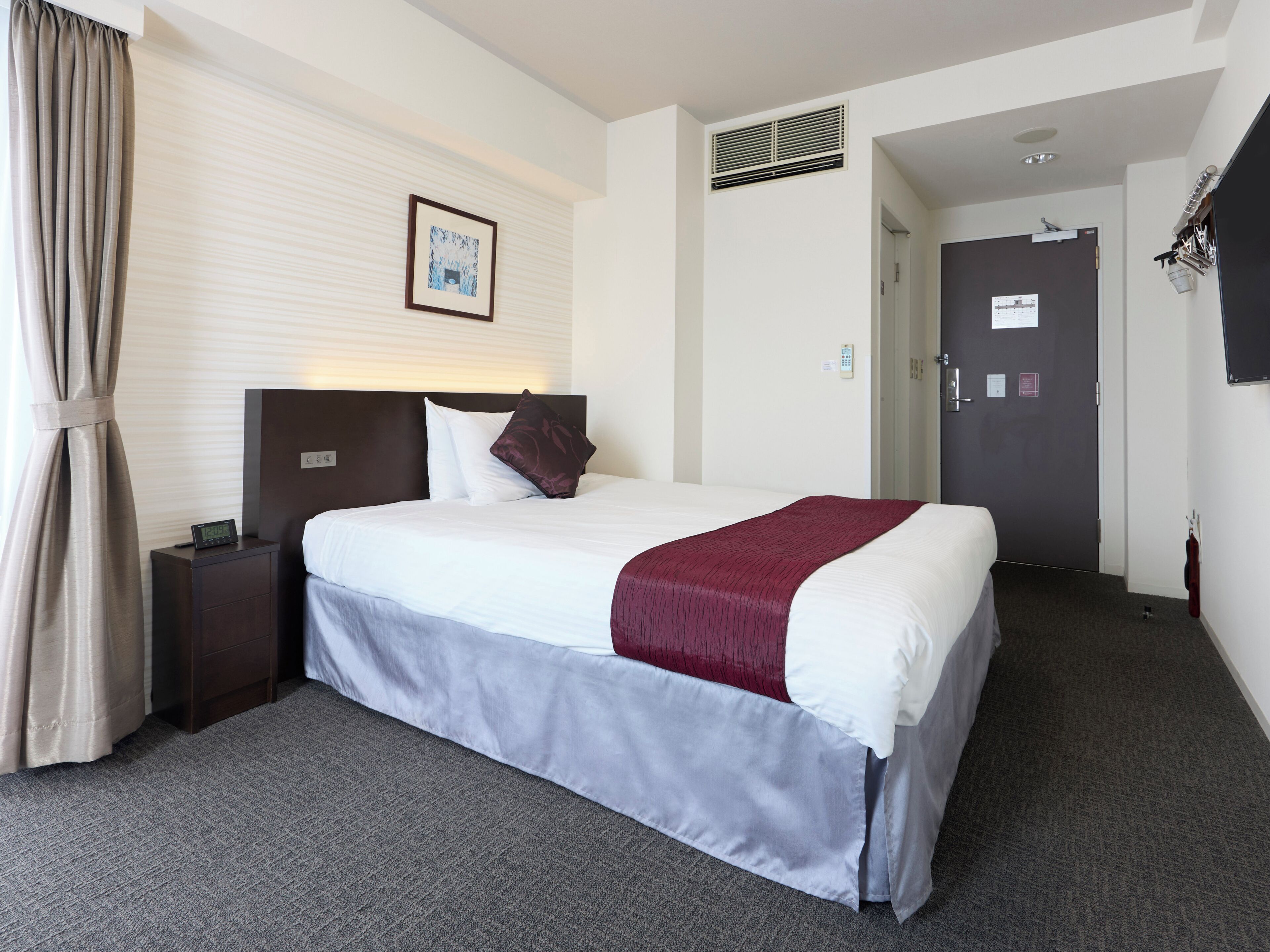 Quarto de hotel com cama, mesa de cabeceira, obra de arte na parede, porta e janela com cortinas.