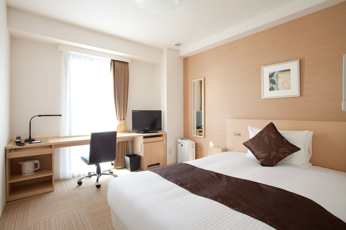 Hotellihuone, jossa on suuri sänky, työpöytä, jossa on tietokone, tuoli, kahvinkeitin ja pieni roskakori.