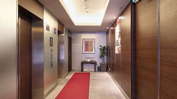 A hotel hallway with a red carpet, elevators, a reception desk, and a framed artwork on the wall.