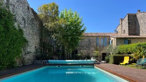 A swimming pool with a wooden deck, stone walls, and a stone building with a tiled roof in the background.