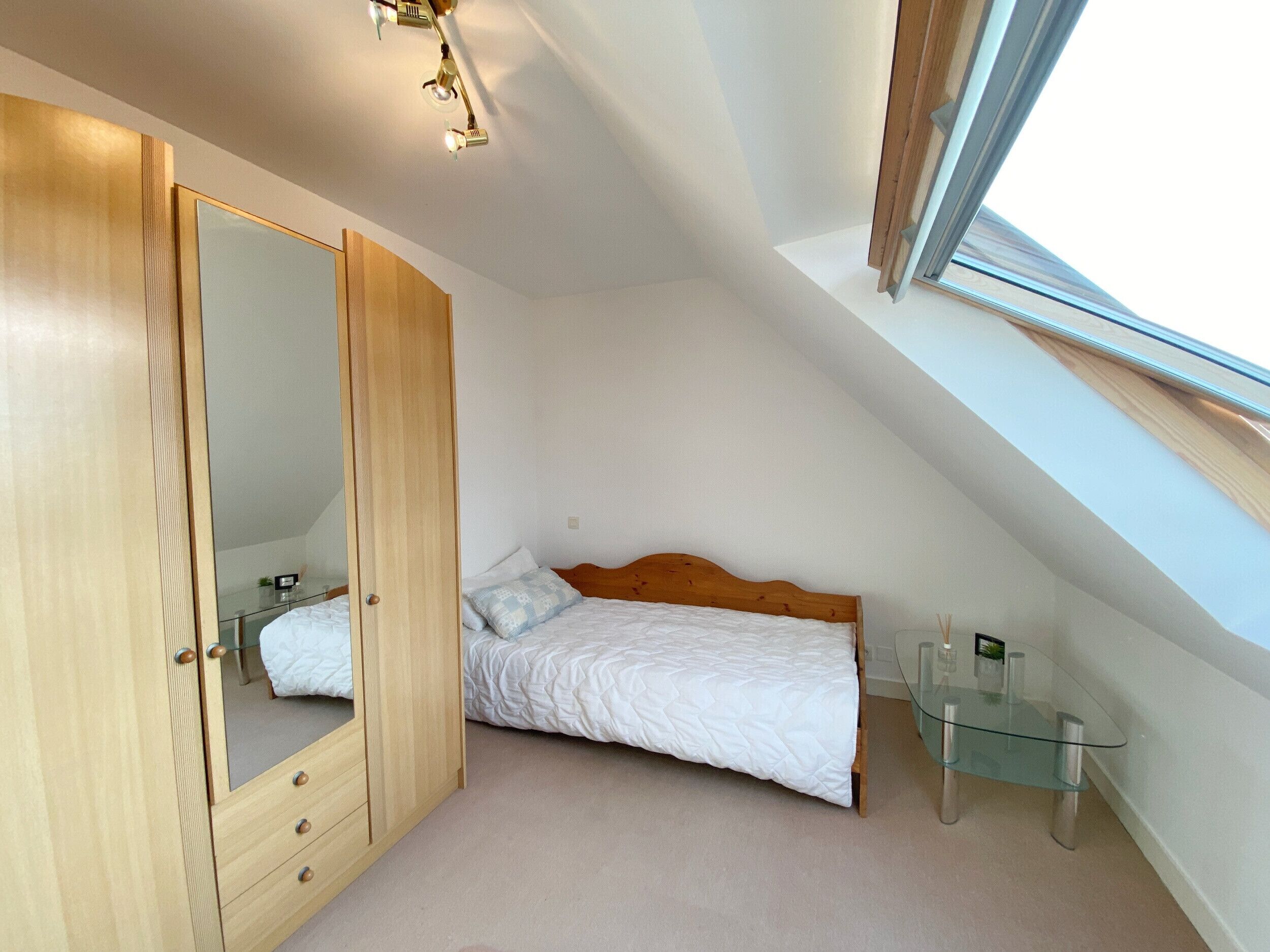 A bedroom with a sloped ceiling, a bed, a wooden wardrobe, a glass table, and a skylight.