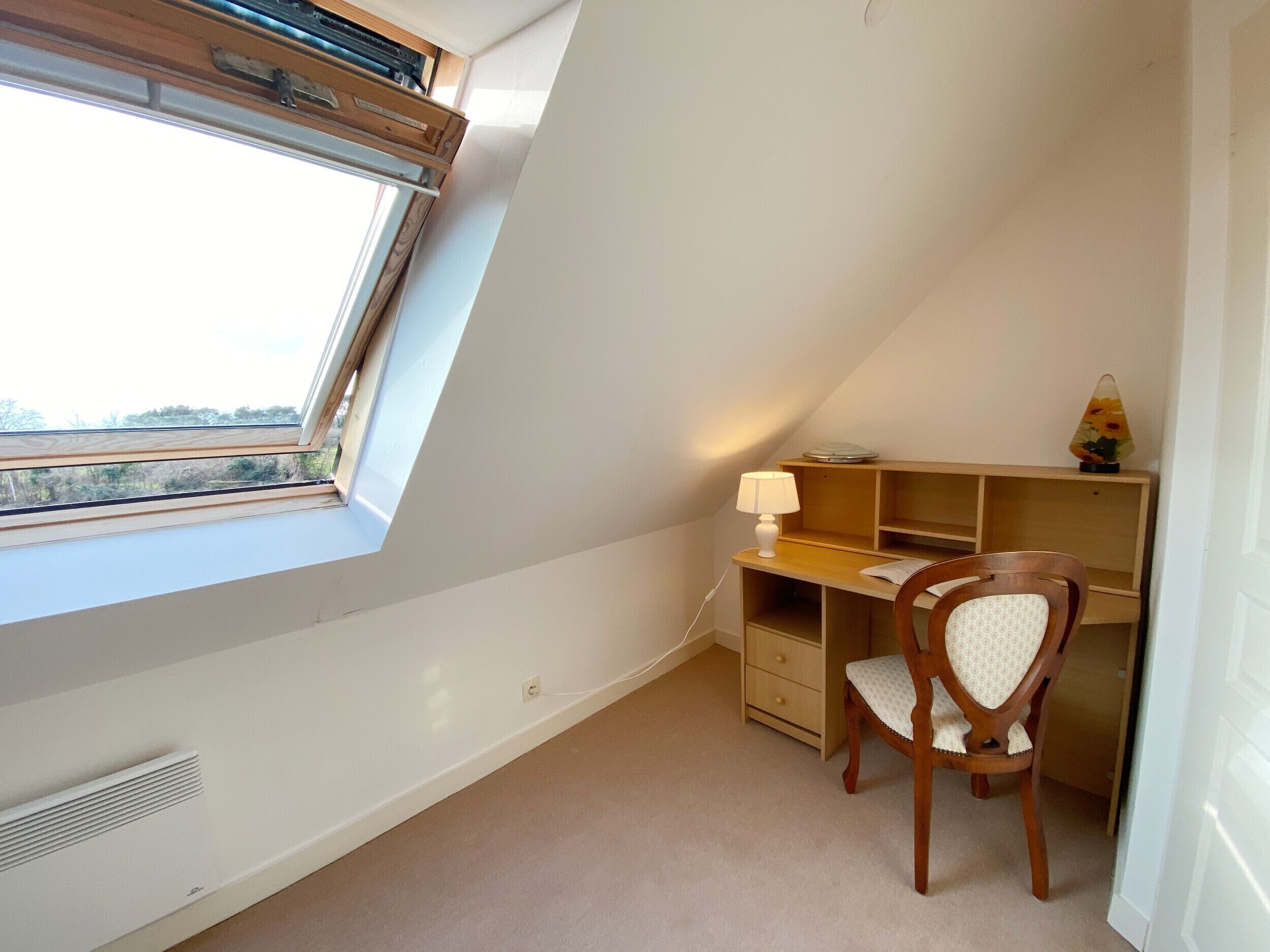 Une pièce avec un plafond en pente, un bureau avec une chaise, une lampe et un radiateur.