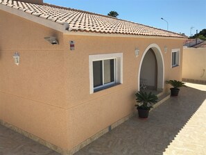 Un edificio con techo de tejas, una ventana pequeña y una puerta con arco. Hay dos plantas en macetas y un farol.