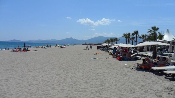 Ein Strand mit Sonnenschirmen und Liegestühlen, Palmen und Bergen im Hintergrund.