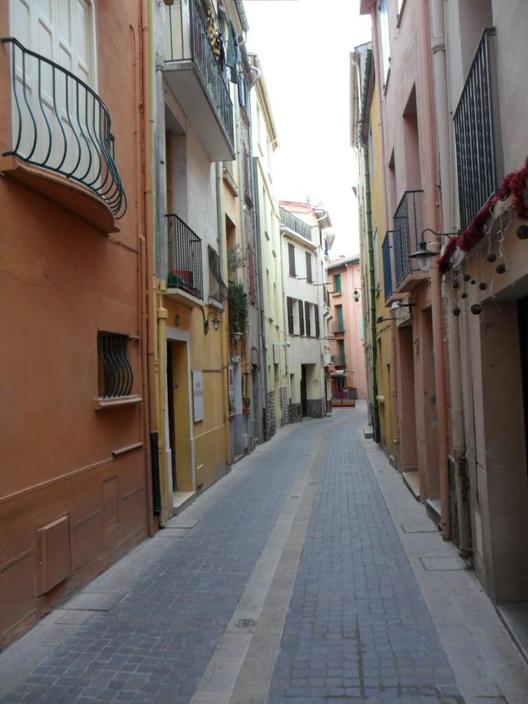 A narrow street lined with multi-story buildings, featuring balconies and a cobblestone path.
