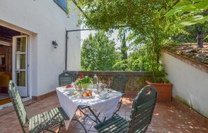 An outdoor patio with a table set for two, surrounded by chairs, and a view of trees and a building in the background.