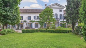 A large, white building with a symmetrical facade, multiple windows with shutters, and a well-maintained garden with hedges and trees.
