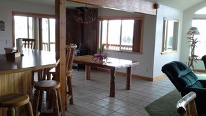 Een ruime eethoek met een houten tafel en stoelen, een keuken op de achtergrond, en grote ramen die natuurlijk licht binnenlaten.