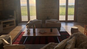 Amplia sala de estar con un ventanal que ofrece vistas a un campo verde. Hay un sofá, una mesa de centro con una botella y una vela, y un televisor sobre un mueble.