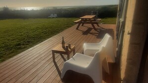 Una terraza de madera con sillas de plástico blanco y una mesa de picnic, con vistas a un campo de césped y una masa de agua.