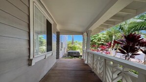 A covered porch with a white railing, a concrete floor, and a view of a residential area with greenery and a parked vehicle. - Hawaii Condo Close to Beach, Golf, Pool and Restaurants (Kapolei)