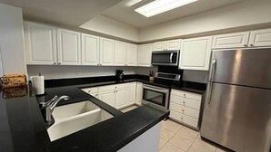 A modern kitchen with white cabinets, a stainless steel refrigerator, a black countertop, and a microwave. - Hawaii Condo Close to Beach, Golf, Pool and Restaurants (Kapolei)