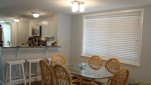 A dining area with a glass table surrounded by wicker chairs, a kitchen with white cabinets and a microwave, and a window with blinds. - Surf Court 48- 1 Block To The Beach! (Hilton Head Island)