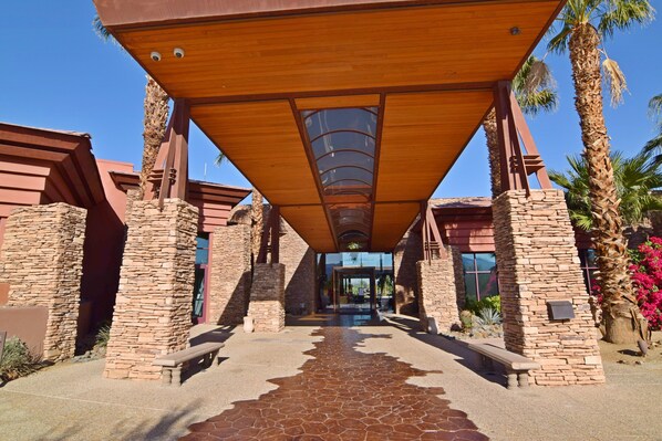 A covered walkway with stone pillars and benches, leading to a building entrance.