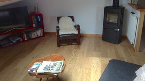 Ett mysigt vardagsrum med trägolv, en fåtölj med en vit pläd, en bokhylla, en tv och en vedkamin.