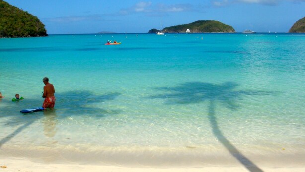 A beach with clear turquoise water, a sandy shore, and a few boats in the distance.