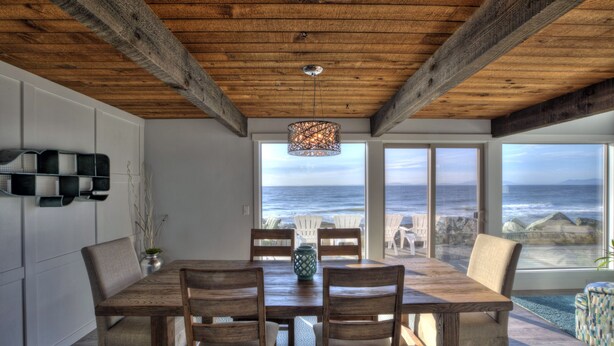 Un espace repas avec une table et des chaises en bois, de grandes fenêtres offrant une vue sur l ’océan et un plafond avec des poutres apparentes.