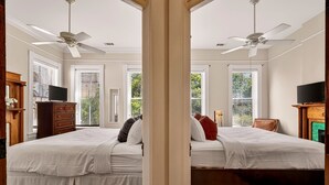 Ein Schlafzimmer mit zwei Betten, Deckenventilatoren, einem Fernseher, einem Kamin und großen Fenstern mit Jalousien.